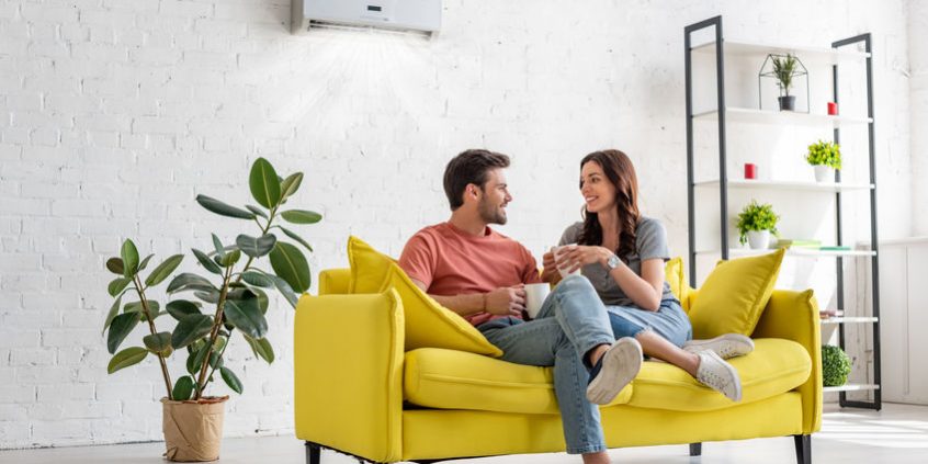 happy young couple talking and holding cups while sitting on yellow sofa under air conditioner at home