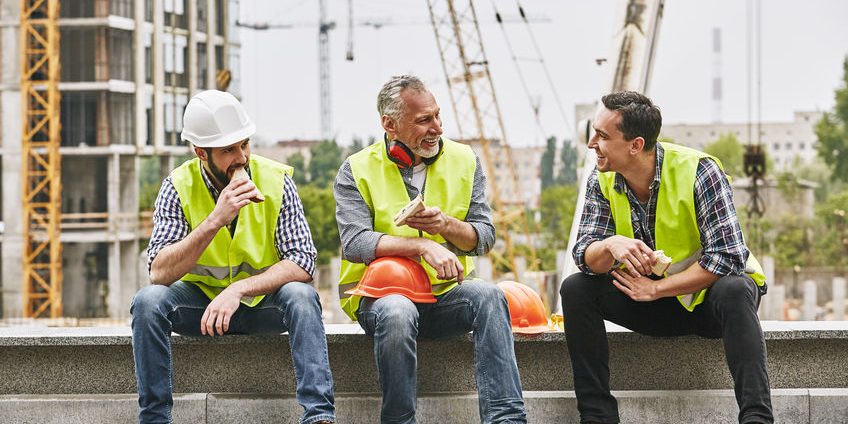 Time for a break. Group of builders in working uniform are eating sandwiches and talking while sitting on stone surface against construction site. Building concept. Lunch concept