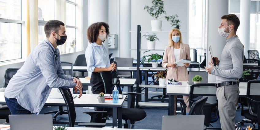 Modern team meeting, group work and social distancing. Manager with tablet speaks with workers in protective masks in interior of modern office with gadgets